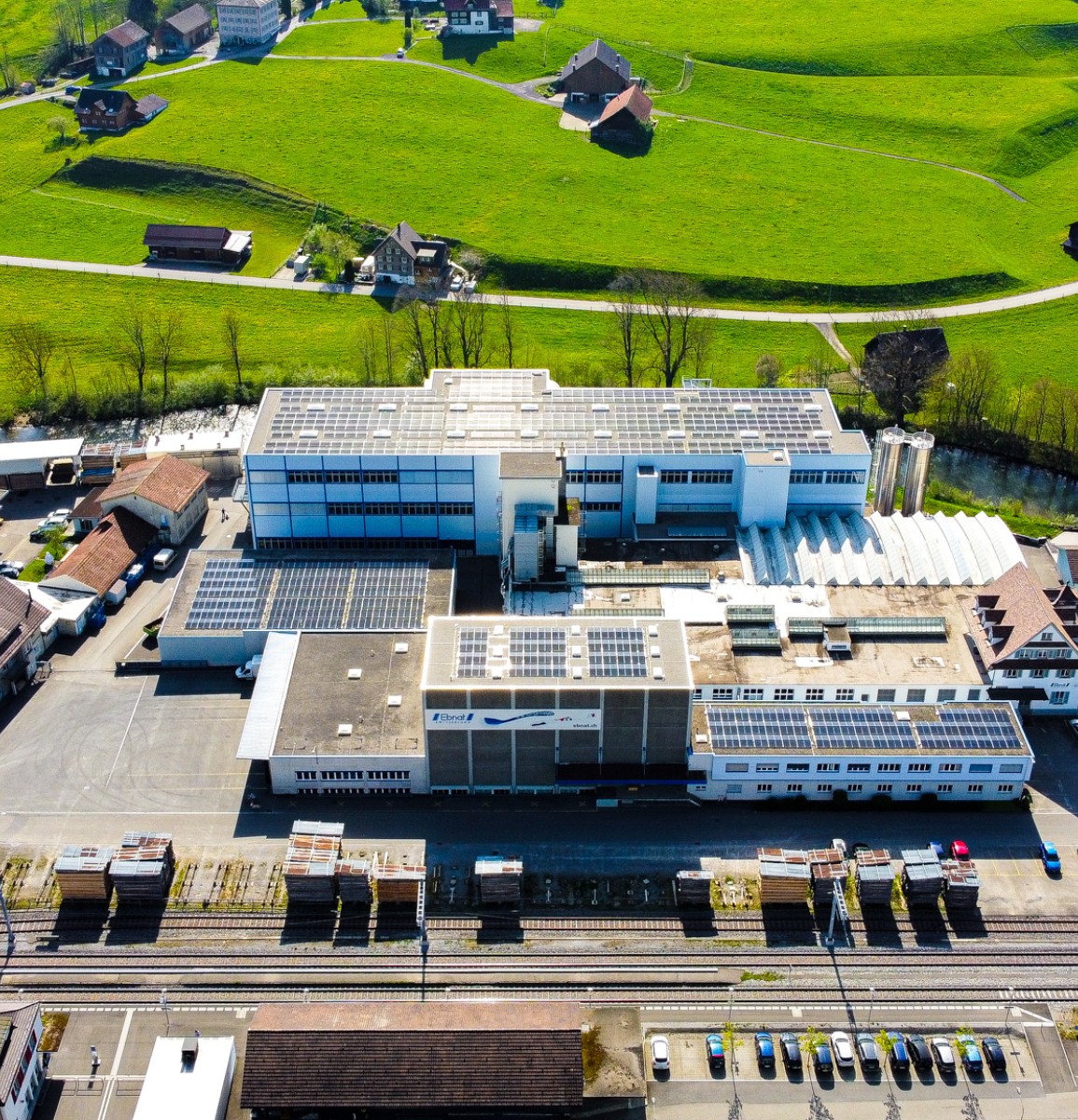 Die Firma Ebnat AG wird von einer Drohne gezeigt. Das ganze Areal der Firma Ebnat AG ist zu sehen. Rundherum sind ältere Häuser. Direkt hinter der Firma befindet sich der Fluss 