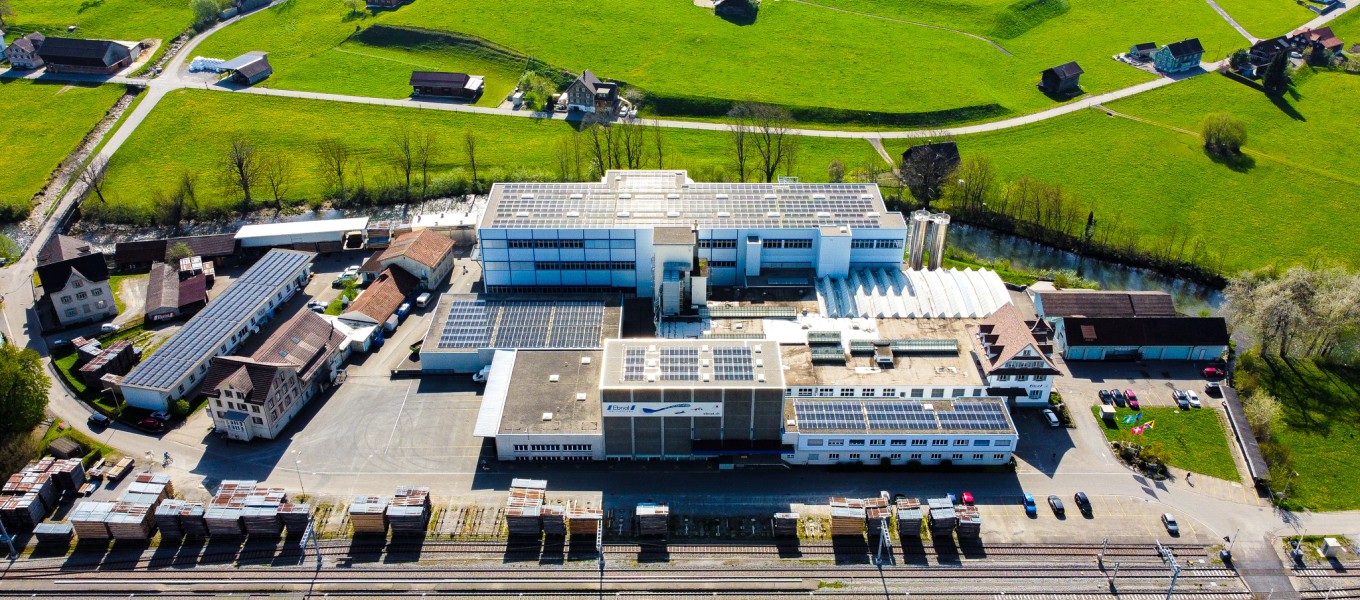 Die Firma Ebnat AG wird von einer Drohne gezeigt. Das ganze Areal der Firma Ebnat AG ist zu sehen. Rundherum sind ältere Häuser. Direkt hinter der Firma befindet sich der Fluss 