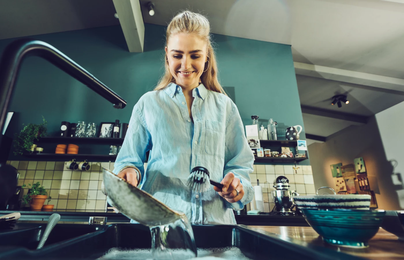 Eine junge Frau in hellblauem Hemd spült lächelnd Geschirr in einer modernen Küche. Sie hält eine schwarze Spülbürste von Ebnat in der Hand und reinigt einen Topf unter fliessendem Wasser. Die Küche im Hintergrund ist stilvoll eingerichtet mit offenen Regalen, Pflanzen und Küchenutensilien.