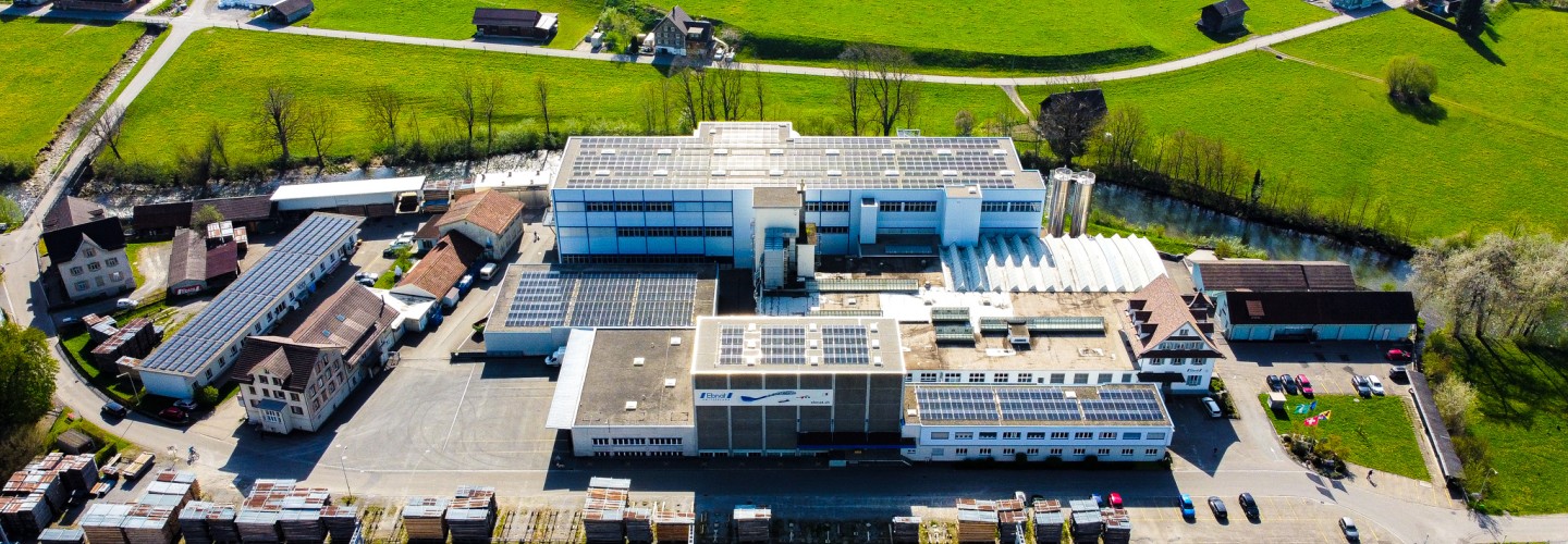 Die Firma Ebnat AG wird von einer Drohne gezeigt. Das ganze Areal der Firma Ebnat AG ist zu sehen. Rundherum sind ältere Häuser. Direkt hinter der Firma befindet sich der Fluss 