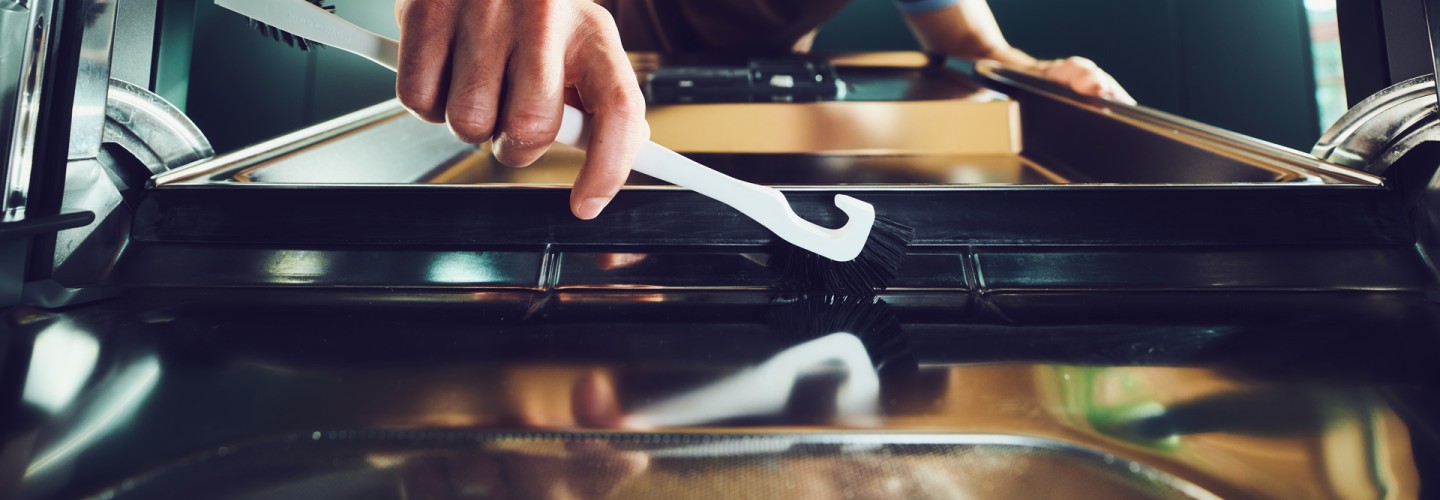 Ein junger Mann mit braunen Locken putzt lächelnd den Geschirrspüler mit einer weissen Universalbürste von der Ebnat AG . Der Mann trägt ein braunes T-Shirt mit hellblauen Rändern. Im Hintergrund ist ein grüner Küchenschrank zu sehen mit einem Blumenstrauss in der Vase drin. Das Zimmer hat Holzwände.