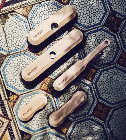 5 Holzbürsten der Firma Ebnat AG liegen auf einem blau/weissen Plattenboden verteilt nebeneinander. Auf der rechten ist es leicht schattig und auf der linken Seite strahlt die Sonne ein. Alle Bürsten sind für die Bodenreinigung.