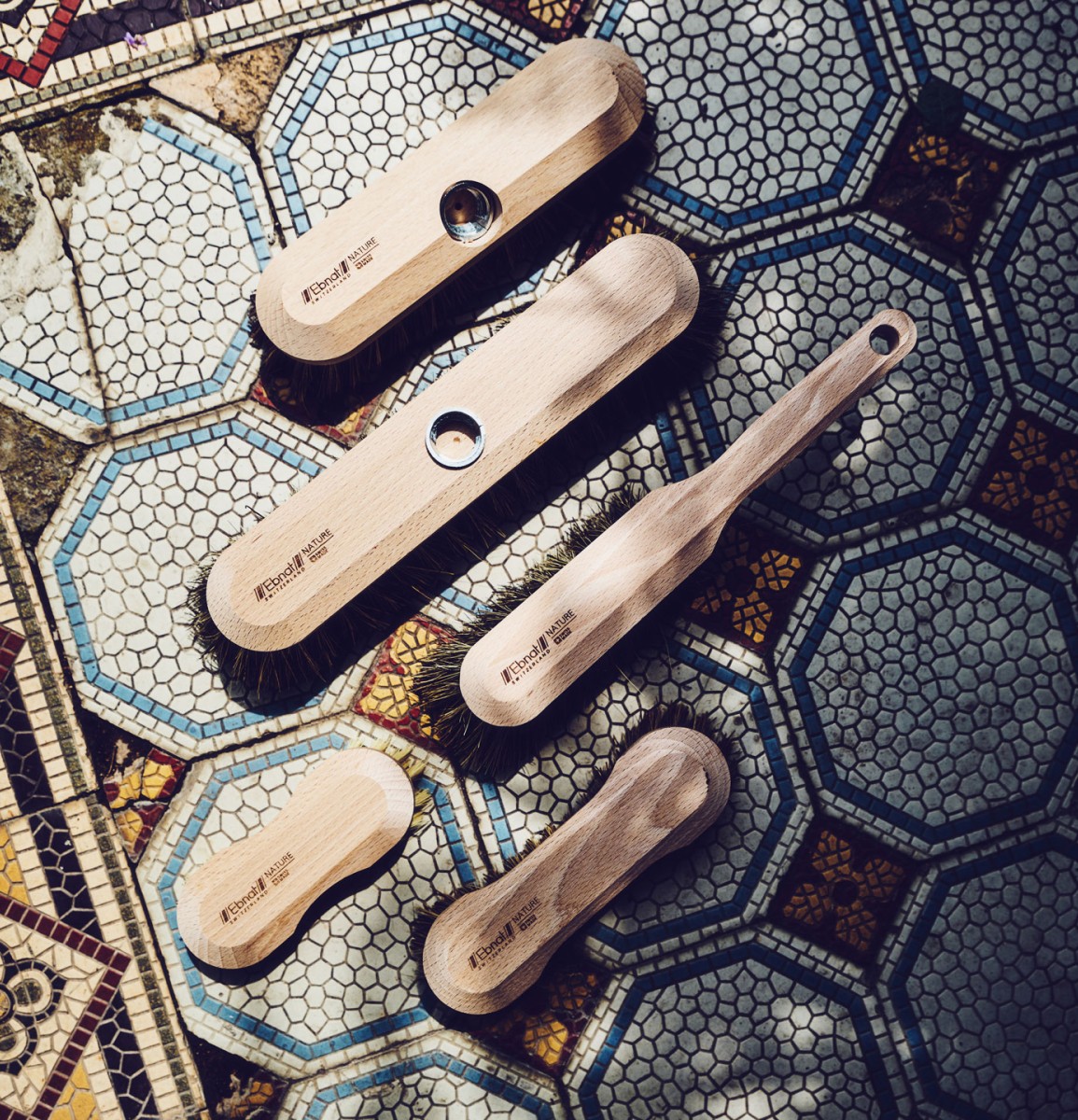 5 Holzbürsten der Firma Ebnat AG liegen auf einem blau/weissen Plattenboden verteilt nebeneinander. Auf der rechten ist es leicht schattig und auf der linken Seite strahlt die Sonne ein. Alle Bürsten sind für die Bodenreinigung.