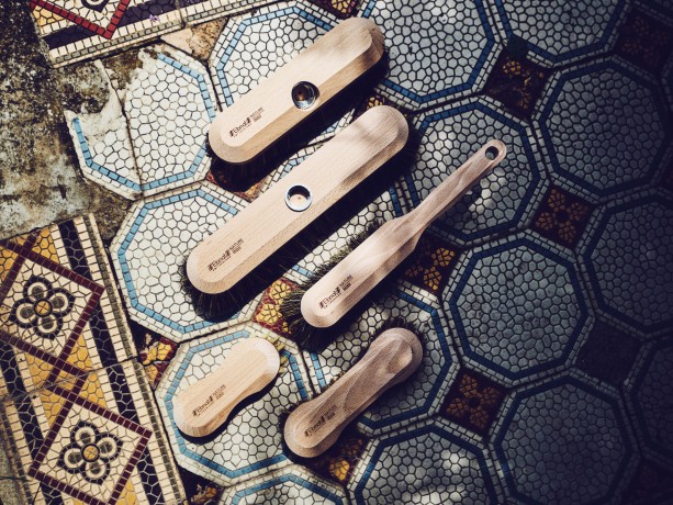 5 Holzbürsten der Firma Ebnat AG liegen auf einem blau/weissen Plattenboden verteilt nebeneinander. Auf der rechten ist es leicht schattig und auf der linken Seite strahlt die Sonne ein. Alle Bürsten sind für die Bodenreinigung.