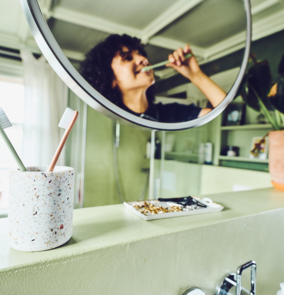 Eine junge Frau mit braunen Locken steht im Badezimmer und putzt sich mit einer grünen Zahnbürste der Ebnat AG die Zähne. Sie blickt sich dabei in einem runden Spiegel an und lächelt. Sie trägt ein dunkelblaues Poloshirt. Links neben dem Spiegel steht ein Zahnputzbecher mit einer grünen und orange farbigen Ebnat AG Zahnbürste. In der Mitte ist eine weisse Ablage mit schmuck darauf. Links vom Spiegel steht eine grosse grüne Pflanze mit einem Terrakotta Topf. Im hinteren Bereich, des hellen Badezimmers, steht ein grünes Regal mit Bildern darin. Nebenan ist eine Dusche aus Glas. Neben der Dusche ist ein geschlossenes Fenster mit einem weissen Vorhang.