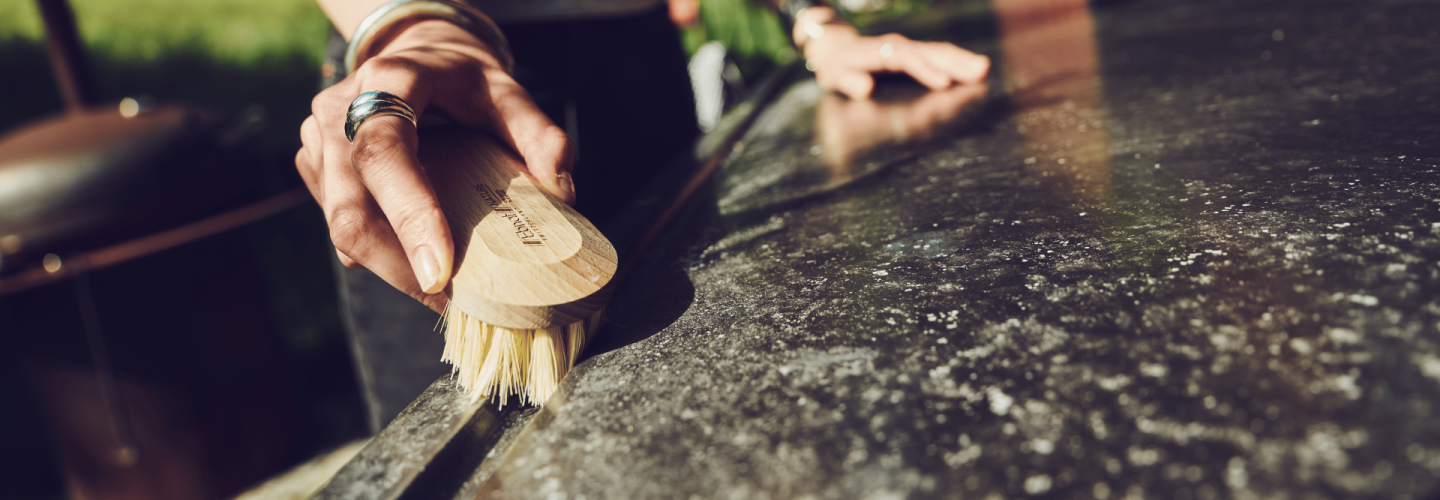 Frau reinigt bei Sonnenlicht eine Outdoor-Oberfläche mit einer Holz-Handbürste von Ebnat; im Hintergrund Natur und Hausfassade mit Pflanzen.