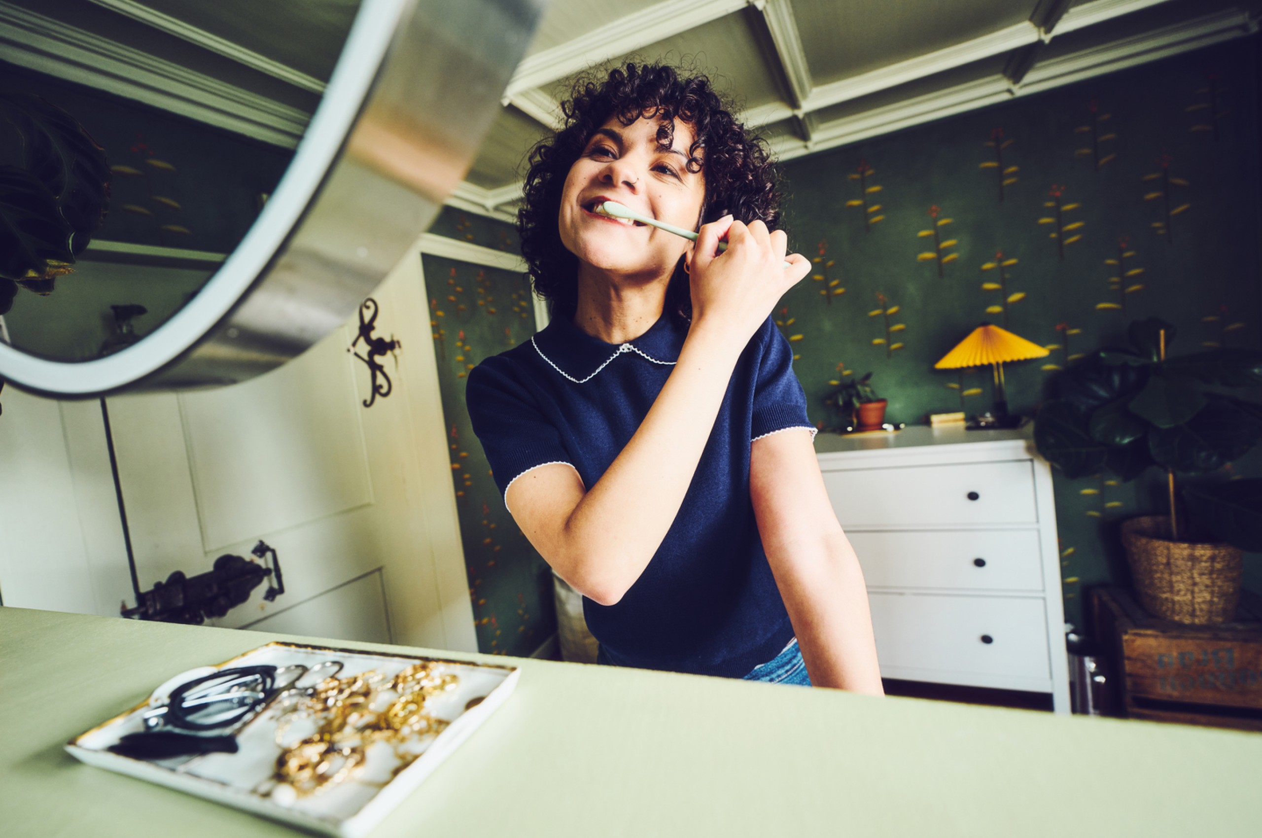 Eine junge Frau mit lockigem Haar putzt sich gut gelaunt die Zähne mit einer Zahnbürste von Ebnat vor einem runden Spiegel. Sie steht in einem liebevoll eingerichteten Raum mit grün gemusterter Tapete. Auf dem Tisch vor ihr liegt Schmuck und eine Brille, im Hintergrund sind eine Kommode, eine gelbe Lampe und eine grosse Zimmerpflanze zu sehen.