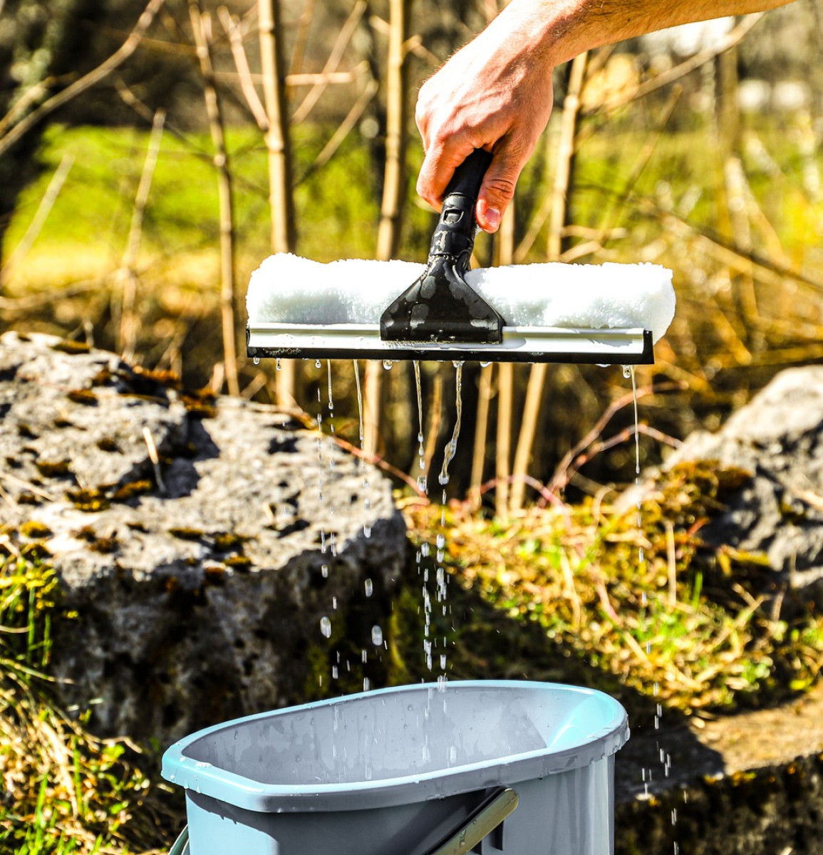 Ein schwarzer Fensterwischer Kombi der Firma Ebnat AG wird aus einem blauen Eimer voll Wasser, von einer männlichen Hand, gezogen. Der Eimer steht in der Natur und wird von der Sonne bestrahlt. Im Hintergrund hat es Steine, Bäume und eine grüne Wiese.