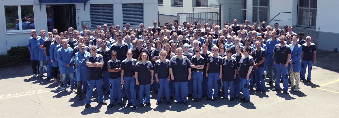 Die Belegschaft der Ebnat AG wird von vorne gezeigt. Sie tragen ihre blauen Arbeitskleider. Die Sonne scheint auf die Mitarbeiter.