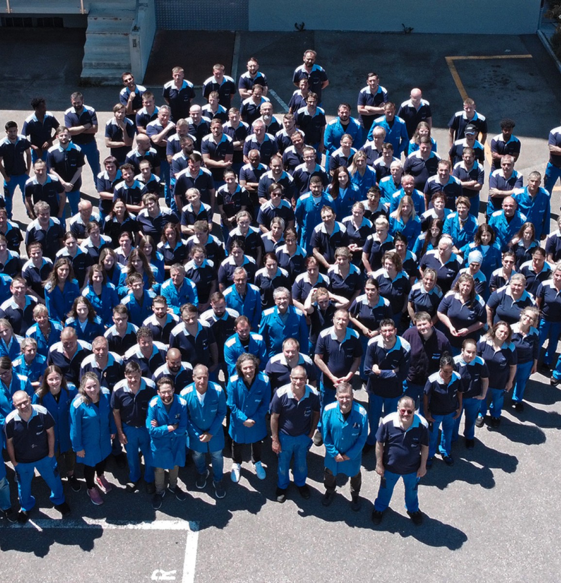 Die gesamte Belegschaft der Ebnat AG ist zu sehen. Es ist eine Aufnahme aus der Vogelperspektive. Die Mitarbeiter lächeln in die Kamera und haben die blauen Arbeitskleider an. Die Sonne scheint auf die Mitarbeiter.