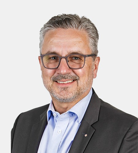 Ein Verkaufsmitarbeiter der Firma Ebnat AG im mittleren Alter lächelt. Er trägt eine graue Brille und hat ein blaues Hemd mit einem schwarze Blazer an. Seine Haare sind grau/weiss. Er hat einen kurzen und weissen Bart.