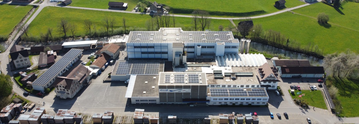 Die Firma Ebnat AG wird von einer Drohne gezeigt. Das ganze Areal der Firma Ebnat AG ist zu sehen. Rundherum sind ältere Häuser. Direkt hinter der Firma befindet sich der Fluss 