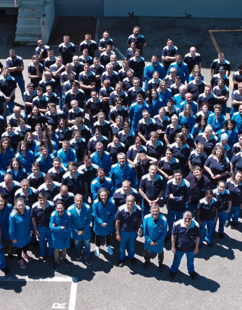 Die gesamte Belegschaft der Ebnat AG ist zu sehen. Es ist eine Aufnahme aus der Vogelperspektive. Die Mitarbeiter lächeln in die Kamera und haben die blauen Arbeitskleider an. Die Sonne scheint auf die Mitarbeiter.