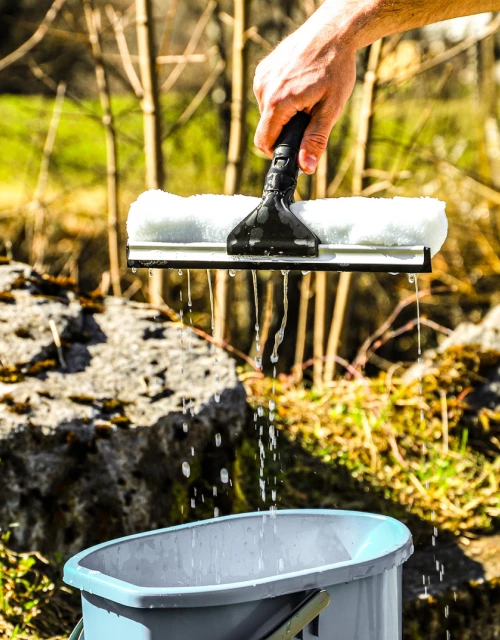 Ein schwarzer Fensterwischer Kombi der Firma Ebnat AG wird aus einem blauen Eimer voll Wasser, von einer männlichen Hand, gezogen. Der Eimer steht in der Natur und wird von der Sonne bestrahlt. Im Hintergrund hat es Steine, Bäume und eine grüne Wiese.