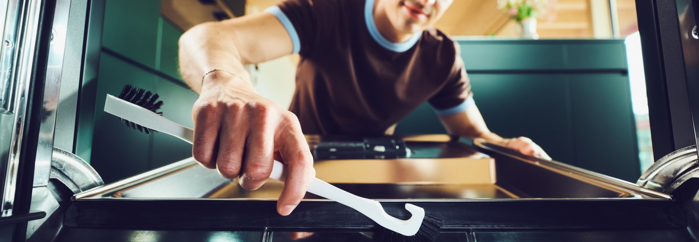 Ein junger Mann mit braunen Locken putzt lächelnd den Geschirrspüler mit einer weissen Universalbürste von der Ebnat AG . Der Mann trägt ein braunes T-Shirt mit hellblauen Rändern. Im Hintergrund ist ein grüner Küchenschrank zu sehen mit einem Blumenstrauss in der Vase drin. Das Zimmer hat Holzwände.