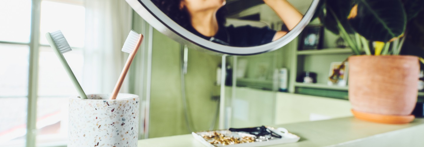 Eine junge Frau mit braunen Locken steht im Badezimmer und putzt sich mit einer grünen Zahnbürste der Ebnat AG die Zähne. Sie blickt sich dabei in einem runden Spiegel an und lächelt. Sie trägt ein dunkelblaues Poloshirt. Links neben dem Spiegel steht ein Zahnputzbecher mit einer grünen und orange farbigen Ebnat AG Zahnbürste. In der Mitte ist eine weisse Ablage mit schmuck darauf. Rechts vom Spiegel steht eine grosse grüne Pflanze mit einem Terrakotta Topf. Im hinteren Bereich, des hellen Badezimmers, steht ein grünes Regal mit Bildern darin. Nebenan ist eine Dusche aus Glas. Neben der Dusche ist ein geschlossenes Fenster mit einem weissen Vorhang.