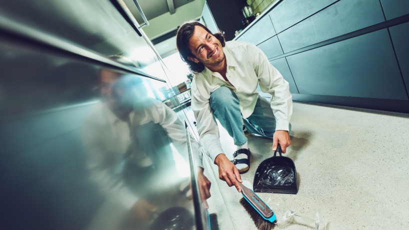 Mann fegt mit einem Handbesen Glasscherben in eine Kehrschaufel auf einem Küchenboden; er lächelt während der Reinigung.