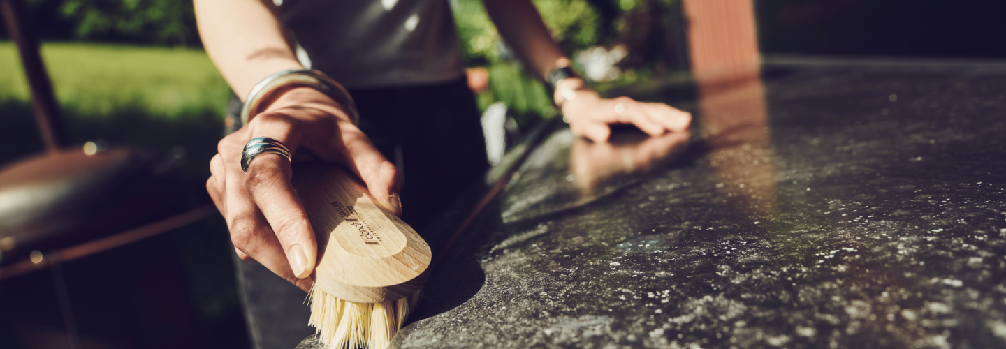 Frau reinigt bei Sonnenlicht eine Outdoor-Oberfläche mit einer Holz-Handbürste von Ebnat; im Hintergrund Natur und Hausfassade mit Pflanzen.