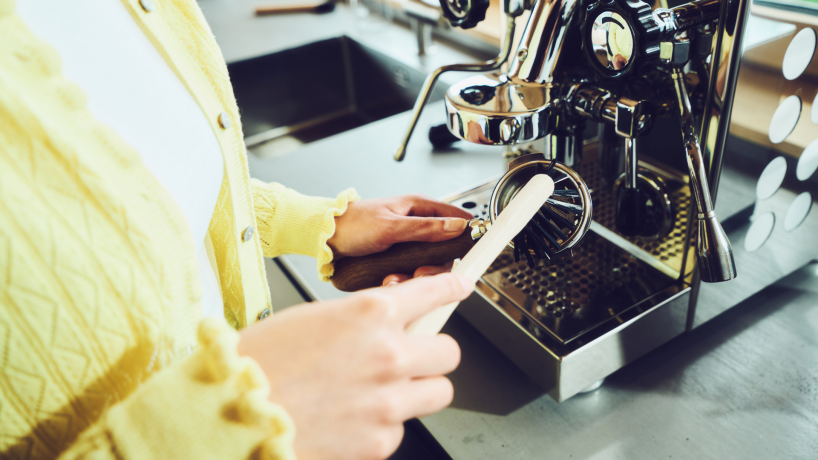 Person in gelbem Pullover reinigt mit einer Holzbürste eine Siebträgermaschine in einer modernen Küche.