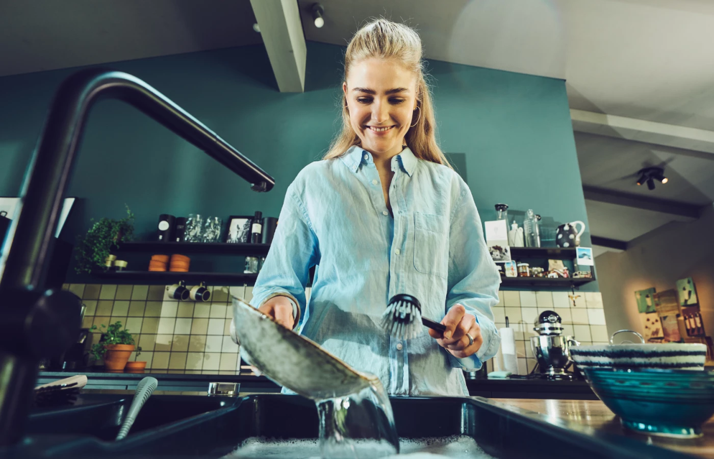 Eine junge Frau in hellblauem Hemd spült lächelnd Geschirr in einer modernen Küche. Sie hält eine schwarze Spülbürste von Ebnat in der Hand und reinigt einen Topf unter fliessendem Wasser. Die Küche im Hintergrund ist stilvoll eingerichtet mit offenen Regalen, Pflanzen und Küchenutensilien.