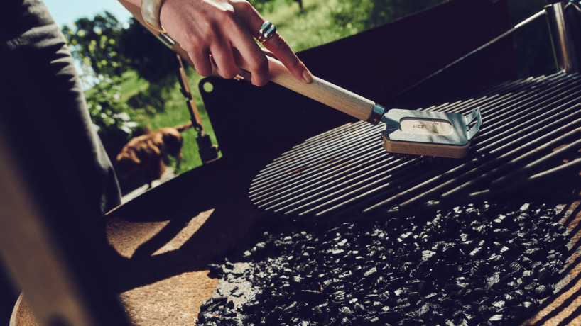 Eine Frau, die einen silbernen Ring und einen Armreifen trägt, putzt mit einer Holzgrillbürste der Ebnat AG einen Grill. Im Hintergrund hat es einen braunen Hund und eine grüne Wiese. Die Sonne strahlt. Der Himmel ist blau.