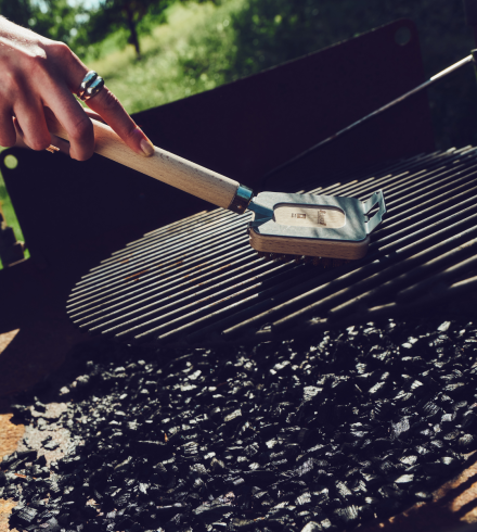 Eine Hand reinigt bei Tageslicht mit einer Grillbürste von Ebnat einen runden Grillrost. Darunter liegen frische Grillkohlen, im Hintergrund ist verschwommenes Grün einer Wiese oder eines Gartens zu erkennen.
